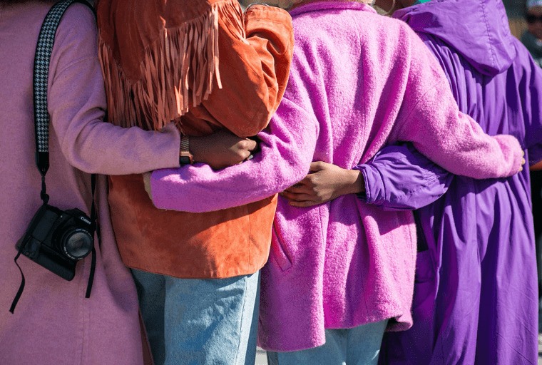 Four people stood with their arms around each other, wearing bright colours